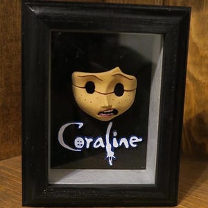 May include: A black shadow box frame containing a sculpted, pale yellow face with hollow black eye sockets, freckles, and an open mouth. Below the face, the word Coraline is written in white script with a blue outline on a black background.