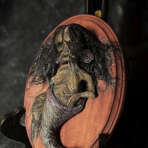 May include: A detailed sculpture of a mermaid with long black hair and a fish tail, mounted on a reddish-brown wooden plaque. The mermaid is depicted with a skeletal face and a purple wrap. The sculpture is displayed on a dark background.