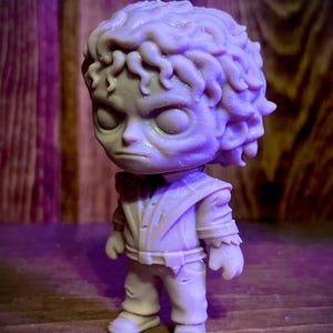 May include: A light gray figurine of a character with a stern expression. The figure has a large head with curly hair and is wearing a detailed outfit. The figurine is standing on a wooden surface.