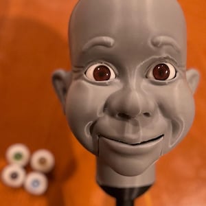 May include: A gray 3D-printed doll head with a smooth finish, featuring brown inset eyes and a hinged jaw line. The head is mounted on a black cylindrical stand. Four additional pairs of interchangeable doll eyes in green, brown, purple, and blue are visible.
