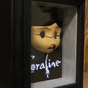 May include: A three-dimensional display featuring a sculpted, pale-skinned face with freckles and an open mouth, mounted inside a black shadow box frame. Below the face, the word Coraline is written in white and blue stylized lettering.