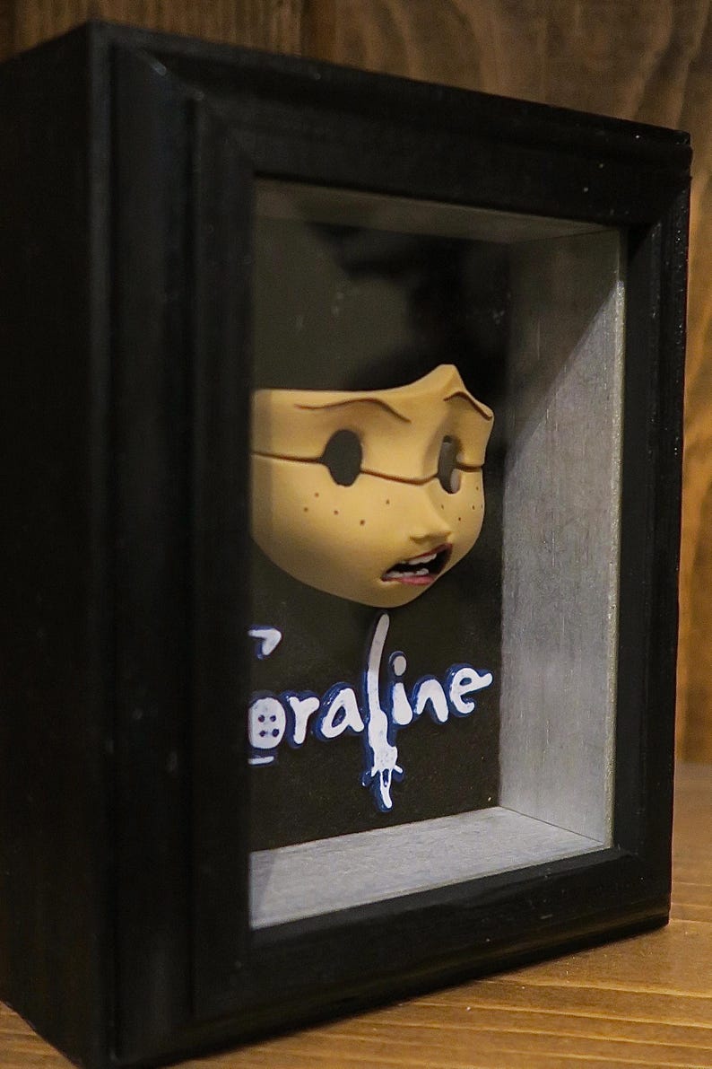 May include: A three-dimensional display featuring a sculpted, pale-skinned face with freckles and an open mouth, mounted inside a black shadow box frame. Below the face, the word Coraline is written in white and blue stylized lettering.