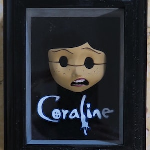 May include: A black framed shadow box containing a sculpted face with dark hollow eyes, freckles, and an open mouth. Below the face, the word Coraline is written in a stylized blue and white font, with a button icon replacing the letter o.