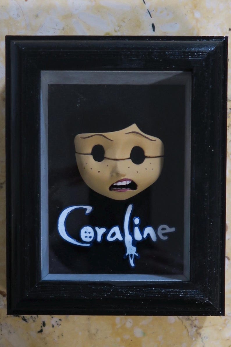 May include: A black framed shadow box containing a sculpted face with dark hollow eyes, freckles, and an open mouth. Below the face, the word Coraline is written in a stylized blue and white font, with a button icon replacing the letter o.