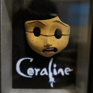May include: A three-dimensional Coraline character face mask mounted in a black frame. The mask is tan with freckles, hollow black eye sockets, and an open mouth showing teeth. The word Coraline is printed in white text below the face.