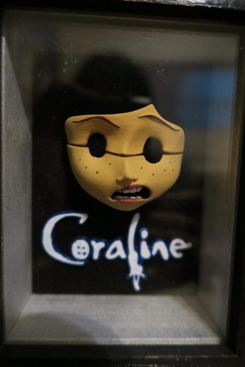 May include: A three-dimensional Coraline character face mask mounted in a black frame. The mask is tan with freckles, hollow black eye sockets, and an open mouth showing teeth. The word Coraline is printed in white text below the face.