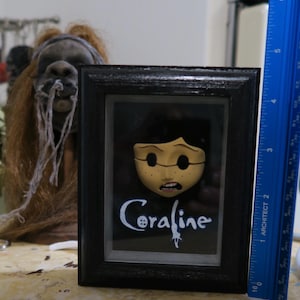 May include: A framed Coraline display featuring a pale, button-eyed character face and the text Coraline in white script. A blue ruler placed vertically next to the frame shows the object is approximately 6 inches tall in this orientation.