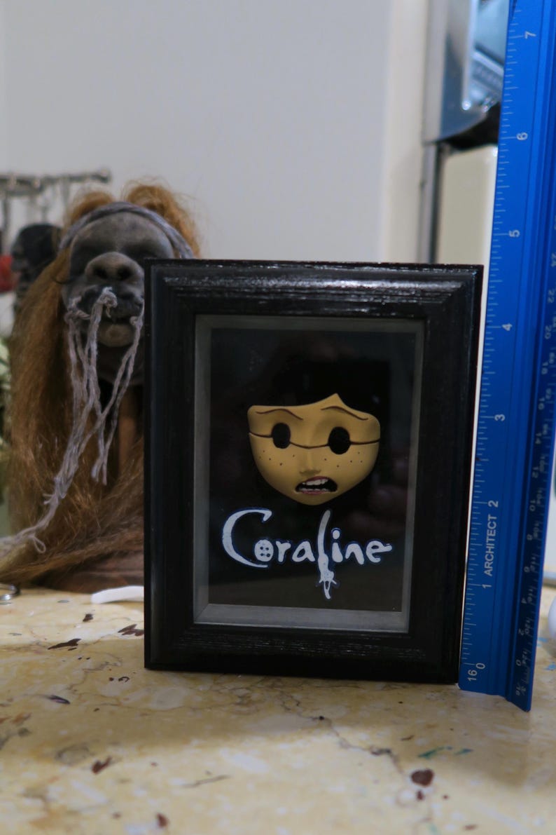 May include: A framed Coraline display featuring a pale, button-eyed character face and the text Coraline in white script. A blue ruler placed vertically next to the frame shows the object is approximately 6 inches tall in this orientation.
