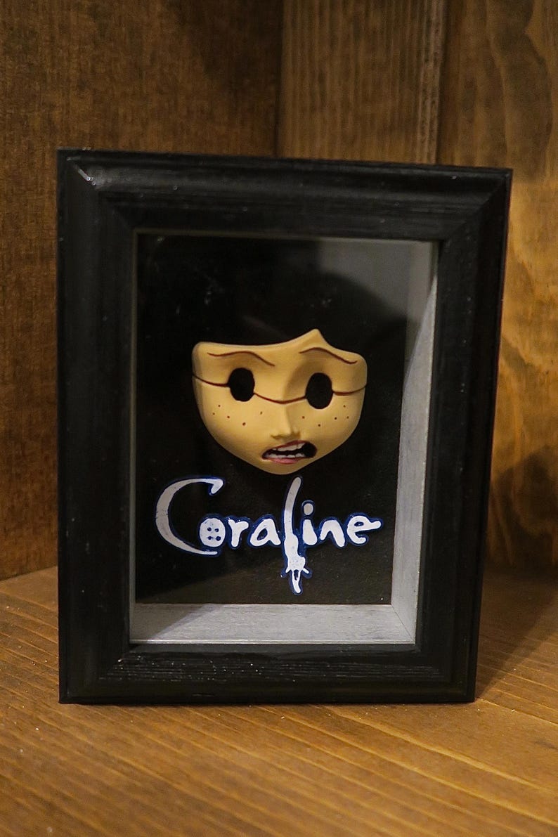 May include: A black shadow box frame containing a sculpted, pale yellow face with hollow black eye sockets, freckles, and an open mouth. Below the face, the word Coraline is written in white script with a blue outline on a black background.