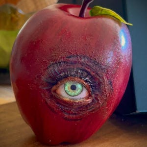 May include: A red apple sculpture with a green eye and long eyelashes. The apple has a stem and a leaf. The eye is detailed with a green iris and a brown outline.