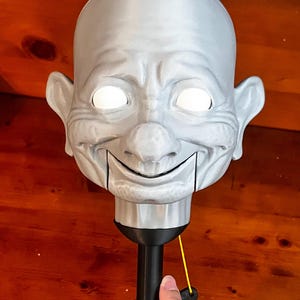 May include: A silver-colored puppet head featuring a bald, expressive face with prominent ears, a large nose, and glowing white eyes. The head is attached to a black cylindrical handle with a thin yellow string mechanism for operating the jaw.