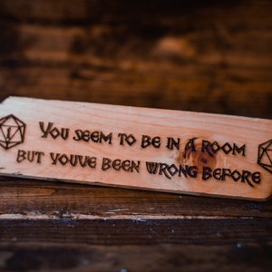 May include: A wooden sign with a burnt-in design of two 20-sided dice and the text "You seem to be in a room but you've been wrong before."