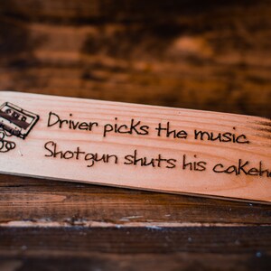 May include: A wooden sign with a burnt wood effect, featuring a cassette tape graphic and the text "Driver picks the music Shotgun shuts his cakehole".
