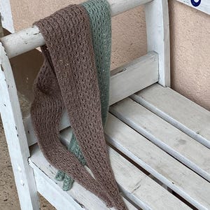 May include: Two knitted scarves, one brown and one green, draped over a white wooden bench. The scarves have a textured pattern. The bench has a seahorse design painted on the side. The background is a light pink wall.