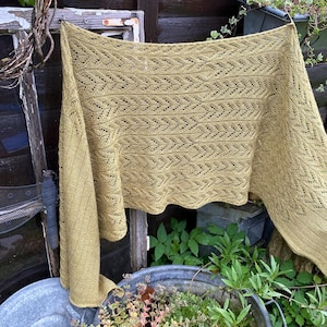 May include: A light olive green shawl with a detailed openwork pattern is displayed outdoors. The shawl is draped over a line, showcasing its intricate design and texture. The background includes greenery and rustic elements.