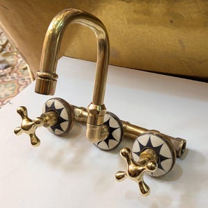 Può includere: Un rubinetto in ottone in stile vintage con un beccuccio curvo e maniglie a forma di croce. Il rubinetto presenta decorativi accenti a stella bianchi e neri sui coperchi delle valvole. L'apparecchio è progettato per un lavandino o una vasca.