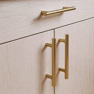 May include: Close-up of a light-colored wooden cabinet with gold-colored hardware. The hardware includes a horizontal bar handle and two vertical T-shaped handles. The cabinet doors have a light wood grain texture.
