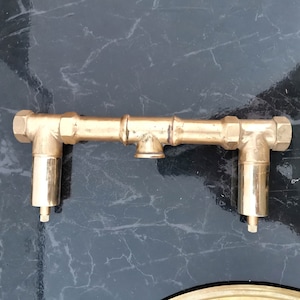 Pu&ograve; includere: Un raccordo per tubi in metallo color oro con due tubi angolari e un tubo centrale. I tubi hanno una finitura lucida.