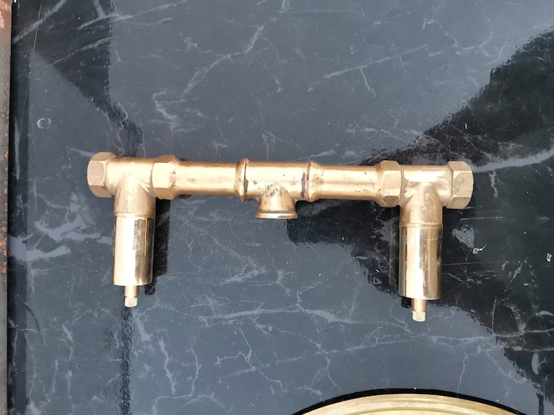 Pu&ograve; includere: Un raccordo per tubi in metallo color oro con due tubi angolari e un tubo centrale. I tubi hanno una finitura lucida.