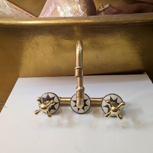May include: A gold-toned faucet with three decorative handles. The handles have a starburst design in black and white. The faucet is mounted on a white surface, with a gold bathtub in the background.