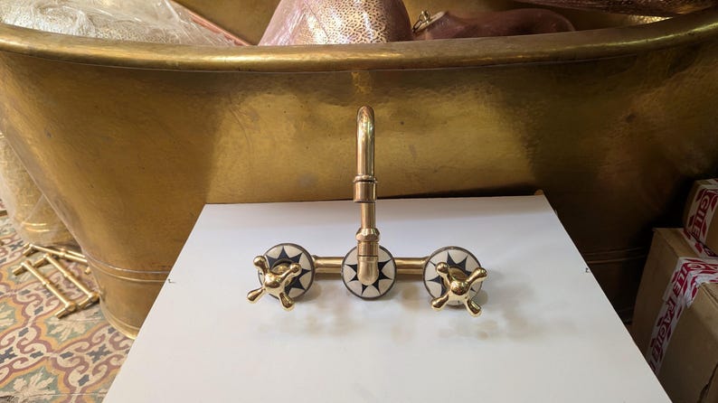 May include: A gold-toned faucet with three decorative handles. The handles have a starburst design in black and white. The faucet is mounted on a white surface, with a gold bathtub in the background.