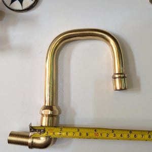 May include: A brass faucet with a curved spout, measuring approximately 7 inches in length, is shown with a measuring tape. Other brass fittings and a decorative circular object with a starburst design are also visible.