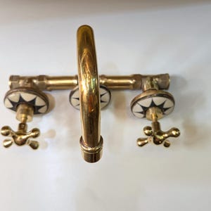 May include: A brass faucet with a curved spout and two handles. The handles are star-shaped and the faucet has white and black decorative accents. The fixture is mounted on a white surface.