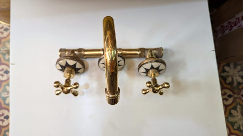 May include: A brass faucet with a curved spout and two handles. The handles are star-shaped and the faucet has white and black decorative accents. The fixture is mounted on a white surface.