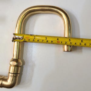 May include: A brass plumbing fitting, bent in a U-shape, is measured with a tape measure. The fitting is approximately 7 inches long. Other brass fittings are visible in the background.