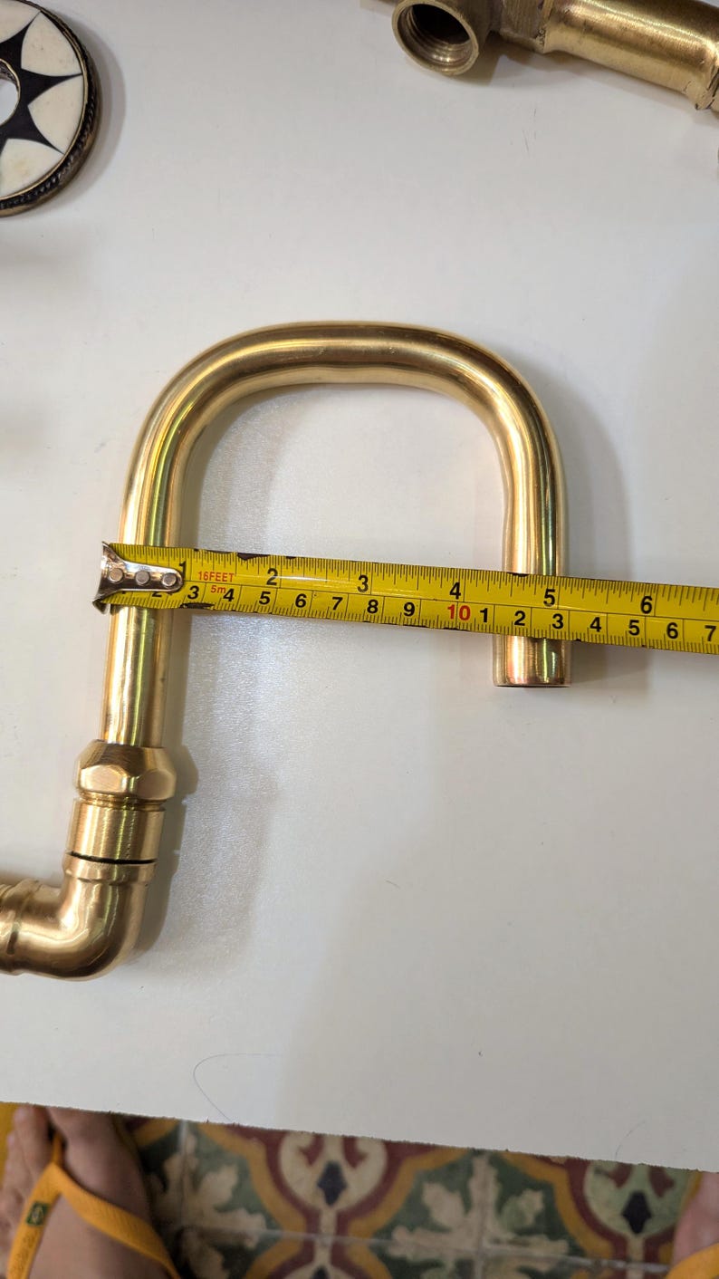 May include: A brass plumbing fitting, bent in a U-shape, is measured with a tape measure. The fitting is approximately 7 inches long. Other brass fittings are visible in the background.
