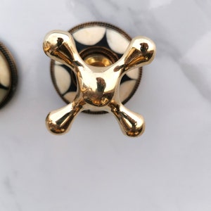 Pu&ograve; includere: Una manopola per rubinetto dorata con una base a motivi bianco e nero. La manopola ha la forma di una croce con estremit&agrave; arrotondate.
