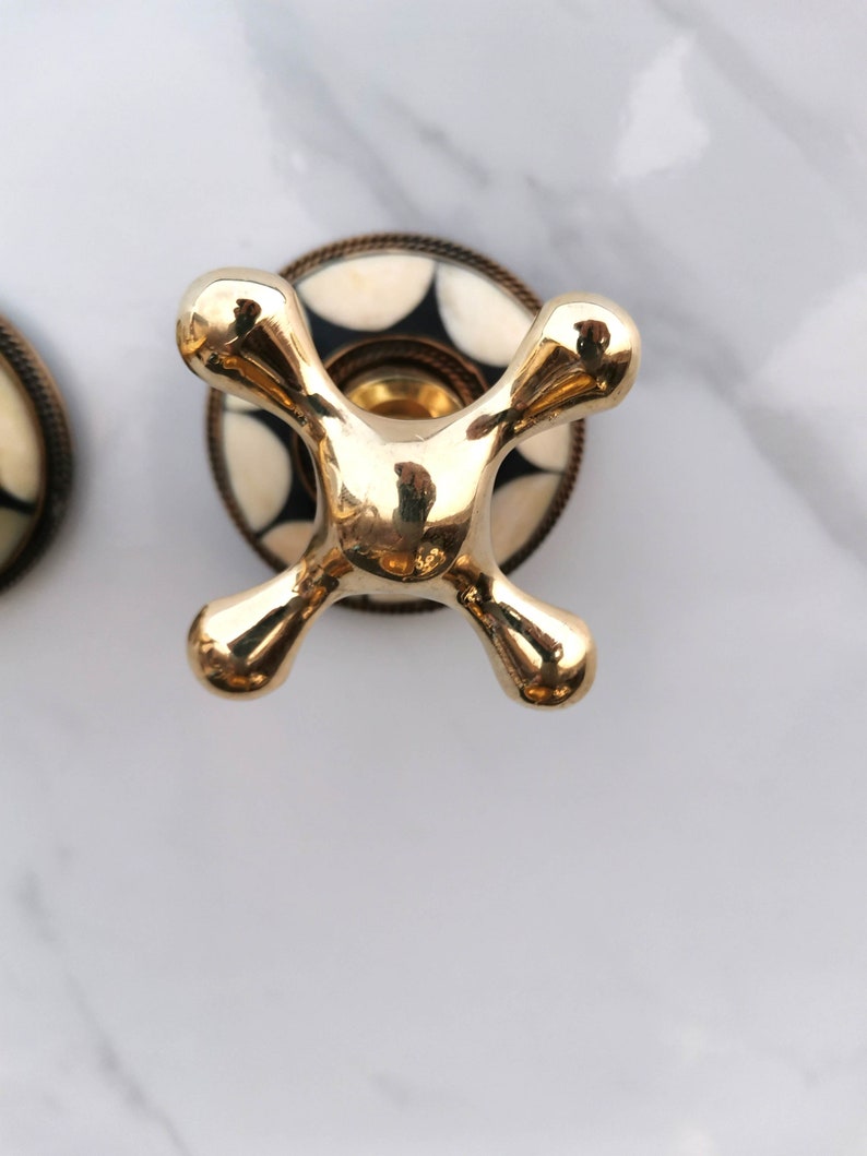 Pu&ograve; includere: Una manopola per rubinetto dorata con una base a motivi bianco e nero. La manopola ha la forma di una croce con estremit&agrave; arrotondate.