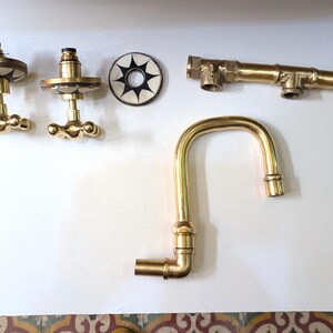 May include: A collection of brass plumbing fixtures, including a faucet, valve handles, and connecting pipes. The faucet has a curved spout. The valve handles have a decorative design. The pieces are arranged on a white surface.