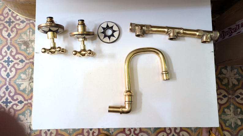 May include: A collection of brass plumbing fixtures, including a faucet, valve handles, and connecting pipes. The faucet has a curved spout. The valve handles have a decorative design. The pieces are arranged on a white surface.