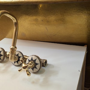 May include: A gold-toned faucet with a curved spout and cross-shaped handles. The faucet has decorative white and black starburst accents. The fixture is likely for a bathroom or kitchen sink.
