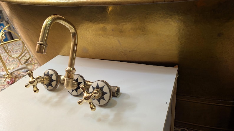 May include: A gold-toned faucet with a curved spout and cross-shaped handles. The faucet has decorative white and black starburst accents. The fixture is likely for a bathroom or kitchen sink.