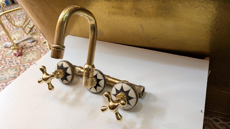 May include: A vintage-style brass faucet with a curved spout and cross-shaped handles. The faucet features decorative white and black starburst accents on the valve covers. The fixture is designed for a sink or tub.