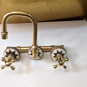 May include: A vintage-style brass faucet with a curved spout and three decorative handles. The handles feature a starburst design in black and white. The faucet is designed for a sink and has a classic, elegant appearance.