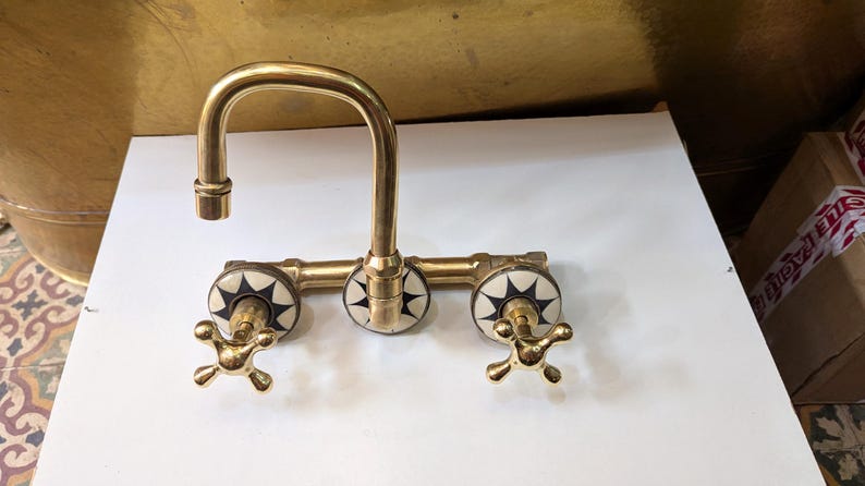 May include: A vintage-style brass faucet with a curved spout and three decorative handles. The handles feature a starburst design in black and white. The faucet is designed for a sink and has a classic, elegant appearance.