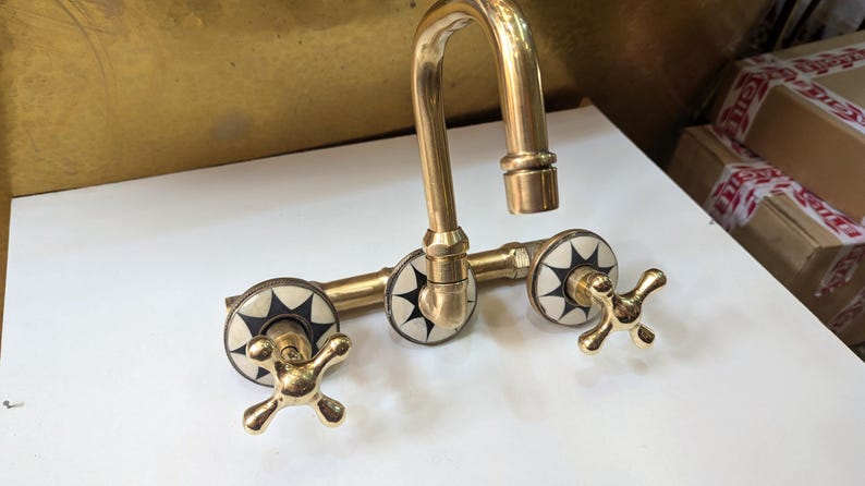May include: A vintage-style brass faucet with a curved spout and cross-shaped handles. The faucet features decorative white and black starburst accents on the handles. The fixture is made of polished brass and is designed for a sink.