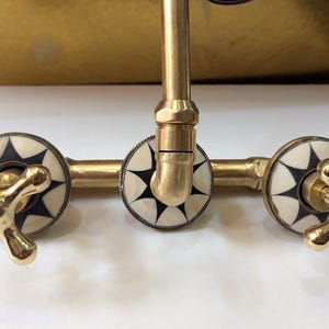 May include: A brass faucet with a cross-shaped handle design. The faucet features decorative white and black starburst accents on the handles and around the base. The fixture has a vintage aesthetic.