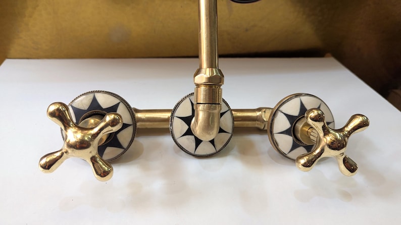 May include: A brass faucet with a cross-shaped handle design. The faucet features decorative white and black starburst accents on the handles and around the base. The fixture has a vintage aesthetic.