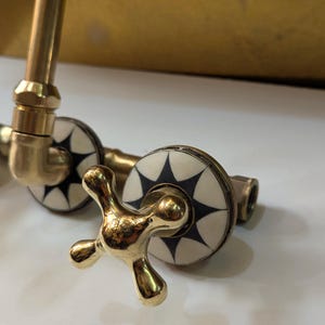 May include: Close-up of a brass faucet with a cross-shaped handle. The faucet has decorative white and black starburst designs on the valve covers. The fixture is set against a white surface.