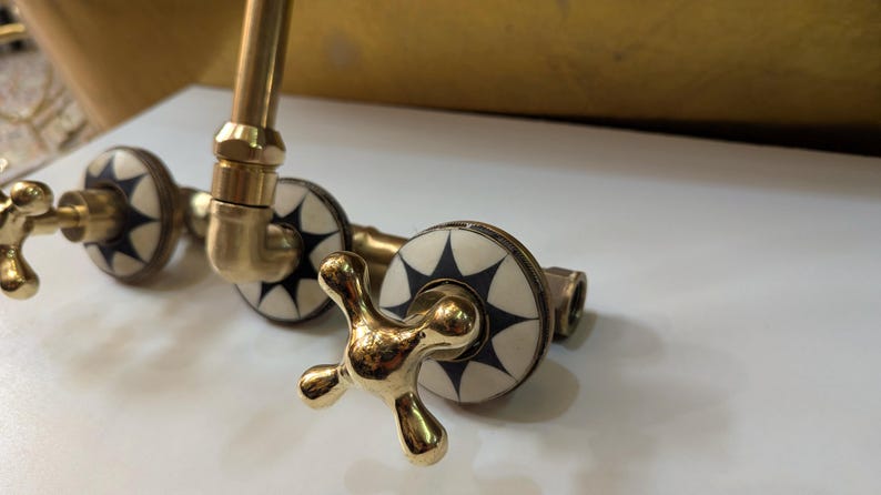 May include: Close-up of a brass faucet with a cross-shaped handle. The faucet has decorative white and black starburst designs on the valve covers. The fixture is set against a white surface.