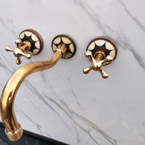 Pu&ograve; includere: Un rubinetto da bagno dorato con tre manopole. Le manopole hanno un design geometrico bianco e nero. Il rubinetto &egrave; montato su una parete di marmo bianco.