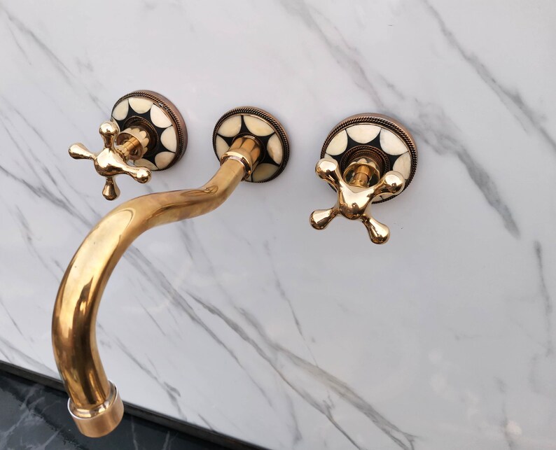Pu&ograve; includere: Un rubinetto da bagno dorato con tre manopole. Le manopole hanno un design geometrico bianco e nero. Il rubinetto &egrave; montato su una parete di marmo bianco.