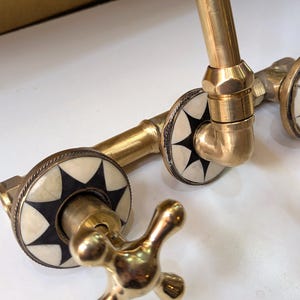 May include: A vintage-style brass faucet with decorative white and black starburst accents. The faucet features cross-shaped handles and is designed for a sink or basin. The brass finish gives it a classic, antique look.