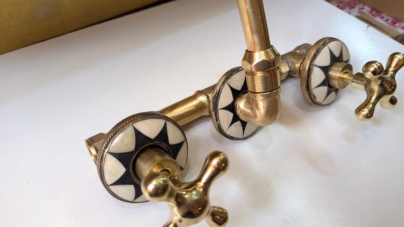 May include: A vintage-style brass faucet with decorative white and black starburst accents. The faucet features cross-shaped handles and is designed for a sink or basin. The brass finish gives it a classic, antique look.