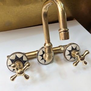 May include: A vintage-style brass faucet with a curved spout and cross-shaped handles. The faucet features decorative white and black starburst accents on the handles. The fixture is made of polished brass and is designed for a sink.