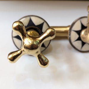 May include: Close-up of a brass faucet with a cross-shaped handle. The faucet has a gold finish and is set against a white background. The faucet's decorative elements include black and white starburst designs.